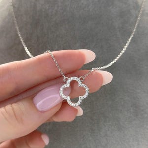 Puede incluir: Un collar de plata con un colgante de trébol de cuatro hojas. El trébol está incrustado con pequeñas piedras preciosas transparentes. El collar se muestra sobre un fondo gris.