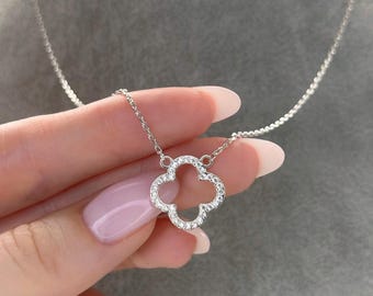 Puede incluir: Un collar de plata con un colgante de trébol de cuatro hojas. El trébol está incrustado con pequeñas piedras preciosas transparentes. El collar se muestra sobre un fondo gris.