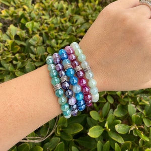 May include: A stack of five colorful gemstone bracelets with silver accents. The bracelets are made of agate, jade, and other semi-precious stones. The bracelets are stacked on a person's wrist.