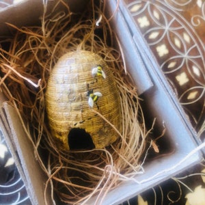 Puede incluir: Una pequeña colmena de cerámica marrón con una superficie texturizada y una pequeña abertura en la parte inferior. Dos pequeñas abejas amarillas y negras están posadas en la colmena.
