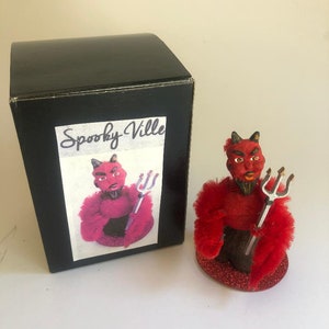 May include: A red devil figurine with a pitchfork, wearing a red fuzzy coat and standing on a red glitter base. The figurine is packaged in a black box with the words "Spooky Ville" printed on the front.