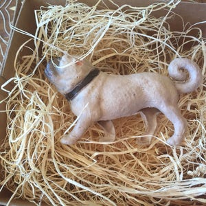 May include: A small, light brown, ceramic dog figurine with a black collar. The figurine is lying in a bed of shredded paper.