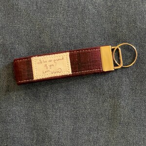 May include: A burgundy fabric keyring with a gold metal ring and a small white fabric tag with the handwritten message "I'm so proud of you! Love, Mom".