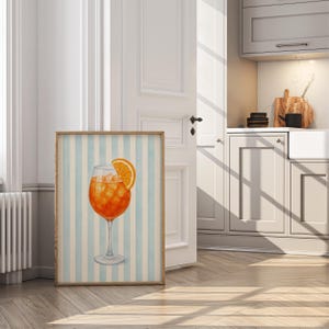 May include: A framed watercolour print depicting an orange cocktail with ice and an orange slice. The drink is in a stemmed glass, set against a light blue and white striped background. The frame is light wood.