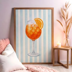 May include: Watercolour print of an Aperol Spritz cocktail in a stemmed glass with an orange slice. The drink is orange with ice cubes. The background is a light blue and white striped pattern, framed in a wooden frame. The artwork is displayed in a room setting.