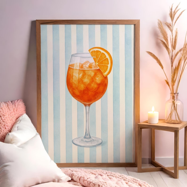 May include: Watercolour print of an Aperol Spritz cocktail in a stemmed glass with an orange slice. The drink is orange with ice cubes. The background is a light blue and white striped pattern, framed in a wooden frame. The artwork is displayed in a room setting.