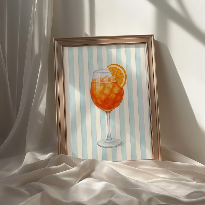 May include: A framed watercolour print of a cocktail. The drink is orange with ice and a slice of orange. The background has blue and white vertical stripes. The frame is gold.