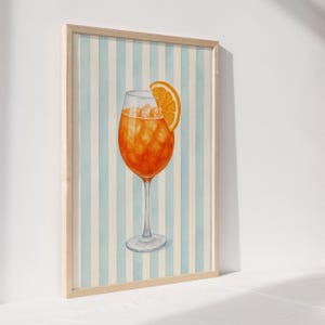 May include: Watercolour print of an Aperol Spritz cocktail in a stemmed glass, garnished with an orange slice. The drink is set against a backdrop of vertical blue and white stripes, framed in a light wood.