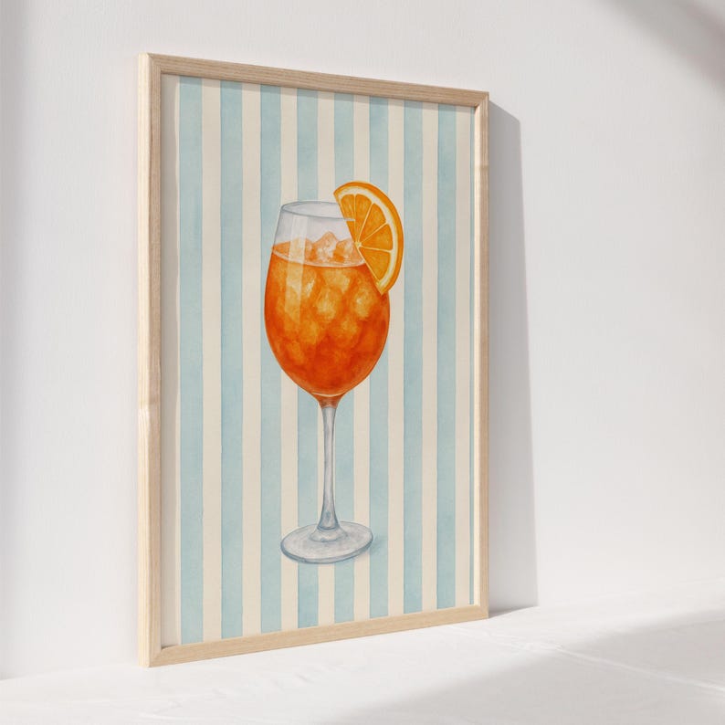 May include: Watercolour print of an Aperol Spritz cocktail in a stemmed glass, garnished with an orange slice. The drink is set against a backdrop of vertical blue and white stripes, framed in a light wood.