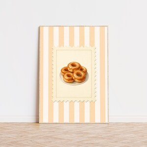 May include: A framed print featuring a stack of glazed donuts on a white plate, set against a cream-colored background with vertical stripes. The donuts are a warm brown color, and the print has a postage stamp-like border.