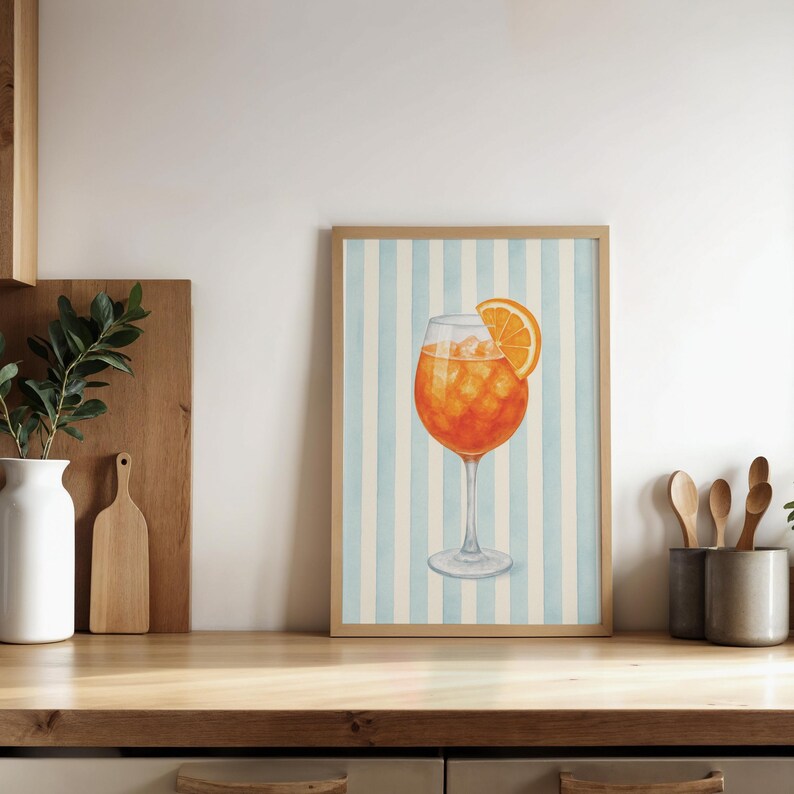 May include: A framed watercolour print of an Aperol Spritz cocktail. The drink is orange with ice and an orange slice, set against a light blue and white striped background. The frame is a light wood colour.
