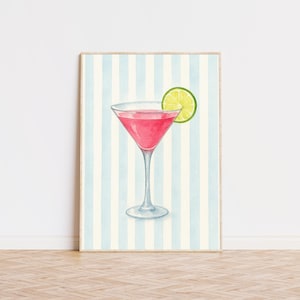 May include: Watercolor print of a pink cocktail in a martini glass with a lime wedge. The artwork is set against a backdrop of vertical blue and white stripes, framed in a light wood. The image evokes a sense of refreshment and leisure.
