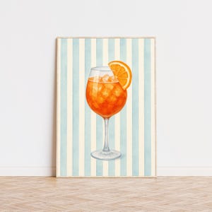 May include: A framed watercolour print of a cocktail in a stemmed glass. The drink is orange, with ice and an orange slice. The background features vertical blue and cream stripes.