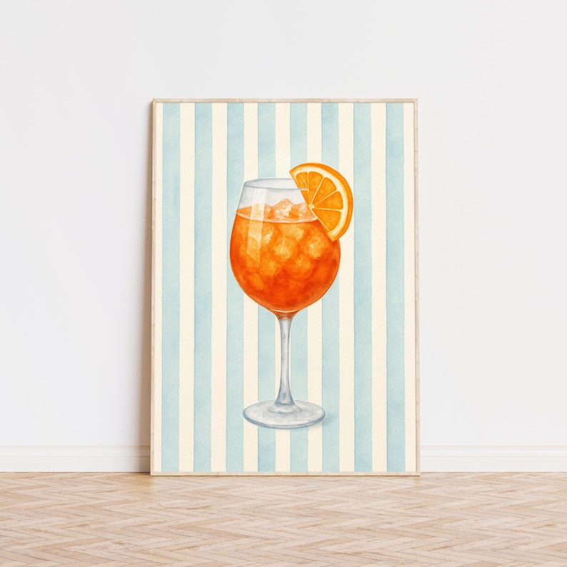 May include: A framed watercolour print of a cocktail in a stemmed glass. The drink is orange, with ice and an orange slice. The background features vertical blue and cream stripes.