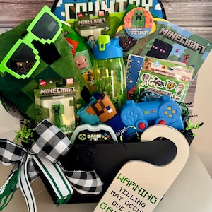 May include: A Minecraft-themed gift basket filled with various items. Includes a backpack, sunglasses, a water bottle, a towel, stickers, a controller, and a door hanger that reads "WARNING YELLING MAY OCCUR DUE TO GAMING."