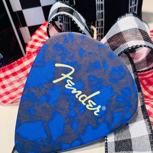 May include: A blue guitar pick with a dark gray pattern and the Fender logo in yellow. The pick is displayed with a red and white checkered ribbon, a black and white checkered ribbon, and a black and white patterned ribbon.
