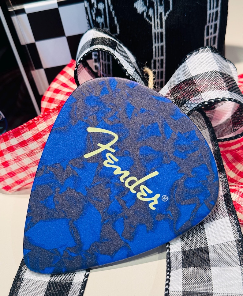 May include: A blue guitar pick with a dark gray pattern and the Fender logo in yellow. The pick is displayed with a red and white checkered ribbon, a black and white checkered ribbon, and a black and white patterned ribbon.