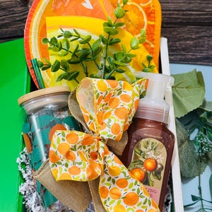 May include: A gift basket with an orange theme. It contains a jar, a bottle of soap, a plate, and a bow with orange and green patterns. The basket is decorated with greenery and sits in a white wooden box.