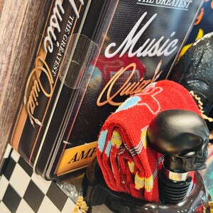 May include: A gift set featuring a black skull-shaped bottle stopper, red socks with a colorful pattern, and a black tin box with "The Greatest Music Quiz" text. The box has a gold and black design.