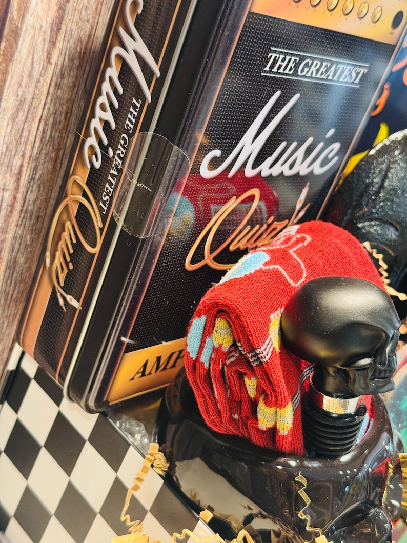 May include: A gift set featuring a black skull-shaped bottle stopper, red socks with a colorful pattern, and a black tin box with "The Greatest Music Quiz" text. The box has a gold and black design.