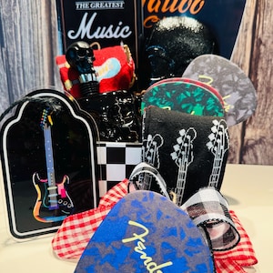 May include: A collection of music-themed items, including guitar picks, a black skull-shaped bottle stopper, and a decorative sign with a guitar illustration. The Fender guitar pick is blue with a gray pattern. The sign reads "The Greatest Music".