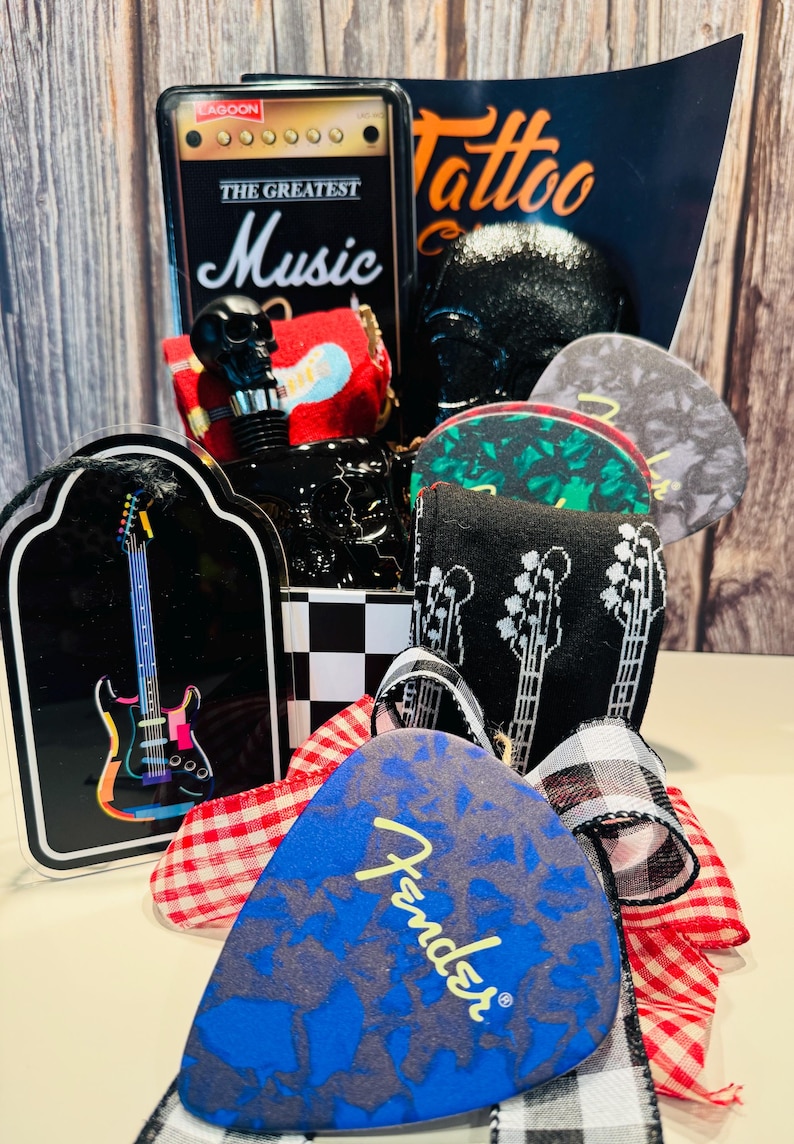 May include: A collection of music-themed items, including guitar picks, a black skull-shaped bottle stopper, and a decorative sign with a guitar illustration. The Fender guitar pick is blue with a gray pattern. The sign reads "The Greatest Music".
