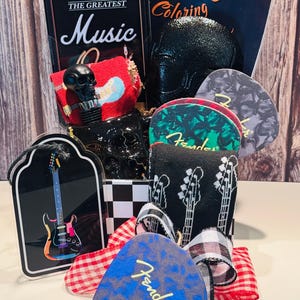 May include: A gift basket with a music theme, featuring a black skull-shaped bottle stopper, guitar picks, a miniature amp, and a coloring book. The guitar picks have the Fender logo and guitar designs. The basket is decorated with ribbons.