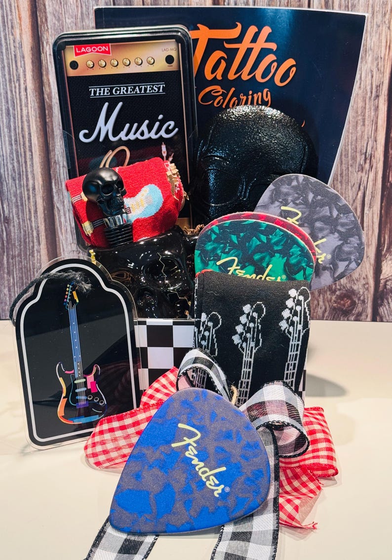 May include: A gift basket with a music theme, featuring a black skull-shaped bottle stopper, guitar picks, a miniature amp, and a coloring book. The guitar picks have the Fender logo and guitar designs. The basket is decorated with ribbons.