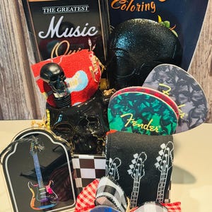 May include: A gift basket with a music theme, featuring a black skull, guitar picks, socks, and a coloring book. The basket includes a tin with "The Greatest Music Quiz" and a red and white checkered bow.