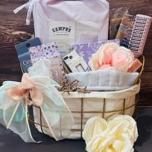 May include: A gold wire basket filled with a light purple Campus Collection sheet set, a floral notebook, a hair comb, cuticle oil, a light blue sleep mask, and pink and white artificial roses, decorated with a large iridescent ribbon bow.