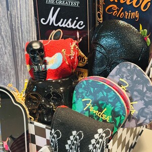 May include: A gift basket with a music theme, featuring a black skull bottle stopper, guitar pick coasters, and a black and white checkered box. Includes a book titled "Tattoo Coloring" and a decorative amplifier.