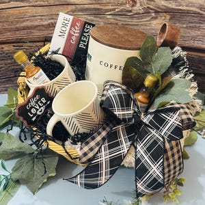 May include: A coffee-themed gift basket with a white coffee canister, two mugs with black geometric patterns, and small bottles of flavoring. The basket includes a decorative bow with a black and white plaid pattern, a sign that says "coffee lover", and a card that says "MORE coffee PLEASE".