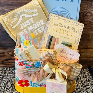 May include: A gift basket featuring a floral container with a large gingham bow, containing a tan pillow with the text Just One More Chapter, a blue PLOT sign, a Once Upon A Pair matching game, a book lover glass, and a sign reading Too Many Plots.