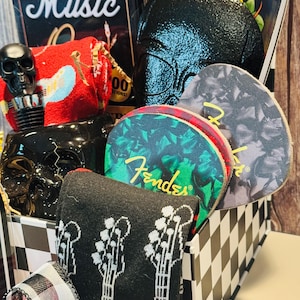 May include: A gift basket with a music theme, featuring a black and white checkered box. Contents include a book titled "The Greatest Music", a black skull bottle stopper, guitar-themed socks, and Fender guitar picks.