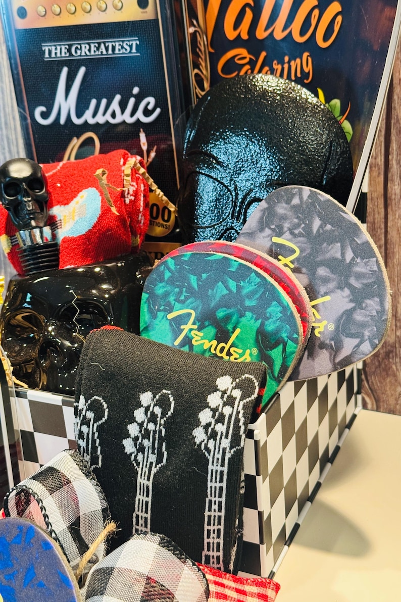 May include: A gift basket with a music theme, featuring a black and white checkered box. Contents include a book titled "The Greatest Music", a black skull bottle stopper, guitar-themed socks, and Fender guitar picks.