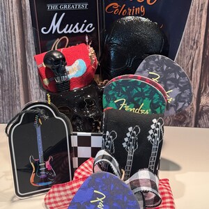 May include: A gift basket with a rock and roll theme. Includes a black skull bottle stopper, guitar picks, socks with guitar graphics, and a miniature amp-shaped box with "The Greatest Music" text. A "Tattoo Coloring" book is also visible.