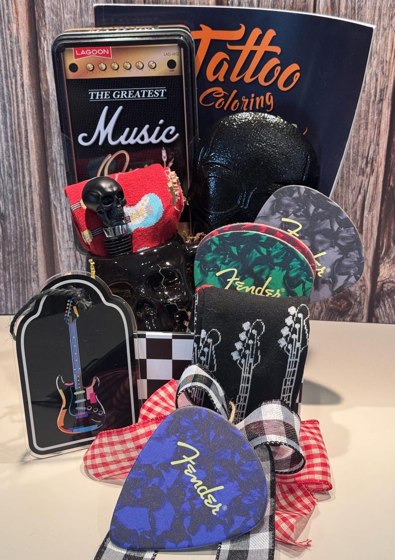 May include: A gift basket with a rock and roll theme. Includes a black skull bottle stopper, guitar picks, socks with guitar graphics, and a miniature amp-shaped box with "The Greatest Music" text. A "Tattoo Coloring" book is also visible.