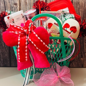 May include: A green basket filled with various items, including a red bow, a glass with a straw, a red pot holder, and a pair of white oven mitts with red floral designs. The basket is decorated with red tinsel and a pink mesh bow.