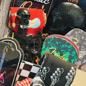 May include: A collection of music-themed items, including a black skull bottle stopper, guitar-themed coasters, and a book titled "Tattoo Coloring." The items are arranged with decorative ribbons and a checkered pattern.
