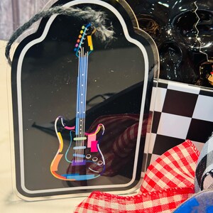May include: A black guitar-shaped acrylic ornament with a colorful electric guitar design. The guitar has a rainbow-colored outline and a blue neck. The ornament is framed with a white border and a black string for hanging.