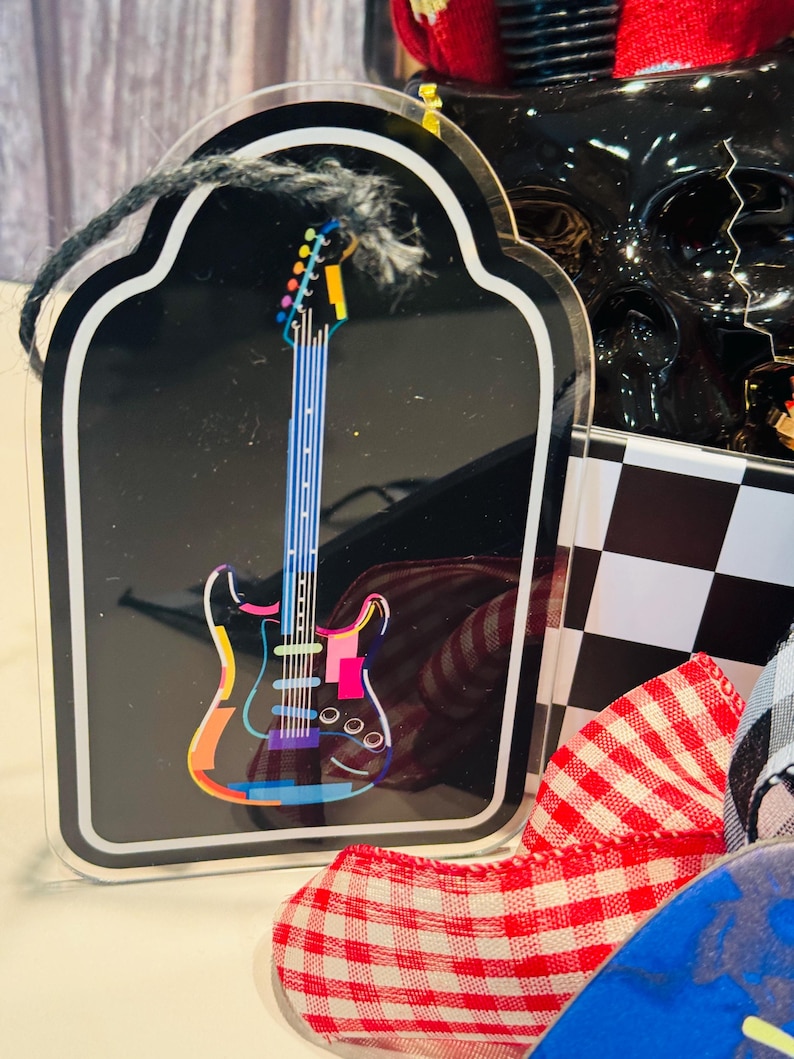 May include: A black guitar-shaped acrylic ornament with a colorful electric guitar design. The guitar has a rainbow-colored outline and a blue neck. The ornament is framed with a white border and a black string for hanging.