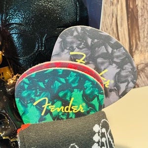May include: A gift set featuring guitar-themed items. Includes guitar picks with the Fender logo, a pair of black socks with a guitar pattern, and a black textured item. The guitar picks have green and gray patterns.