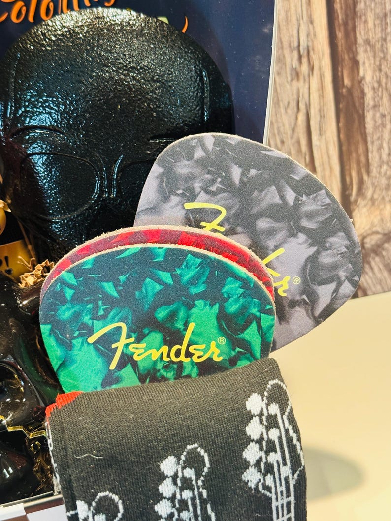 May include: A gift set featuring guitar-themed items. Includes guitar picks with the Fender logo, a pair of black socks with a guitar pattern, and a black textured item. The guitar picks have green and gray patterns.