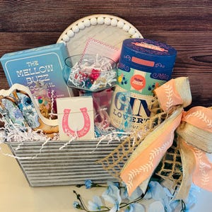 May include: A gift basket containing a variety of items, such as a blue box with "The Mellow Buzz Cocktail" printed on it, a deck of cards with a horseshoe, and a blue tin. The basket is adorned with a peach bow and blue flowers.