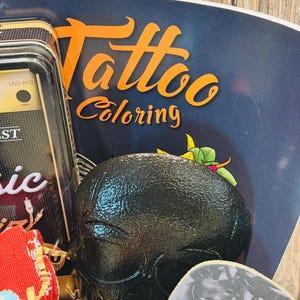May include: A black skull-shaped object is the focal point, surrounded by a dark blue background with the words "Tattoo Coloring" in orange. Other items include a small radio and guitar picks.