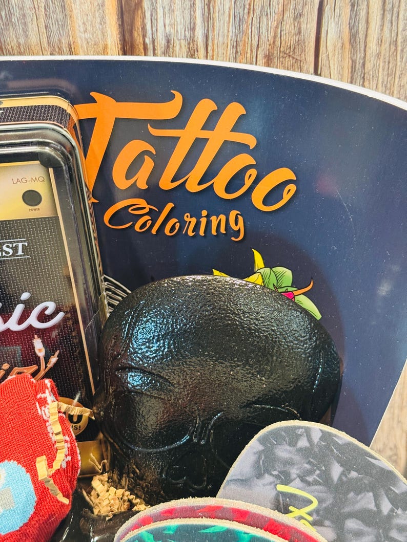 May include: A black skull-shaped object is the focal point, surrounded by a dark blue background with the words "Tattoo Coloring" in orange. Other items include a small radio and guitar picks.