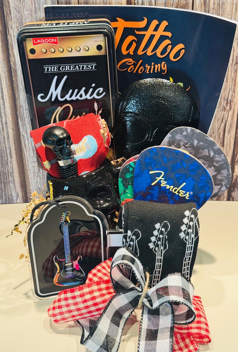 May include: A gift basket with a music theme. Includes a black skull bottle stopper, guitar pick set, guitar-themed socks, a coloring book, and a decorative tin with "The Greatest Music" on it. The basket is tied with a red and black checkered ribbon.