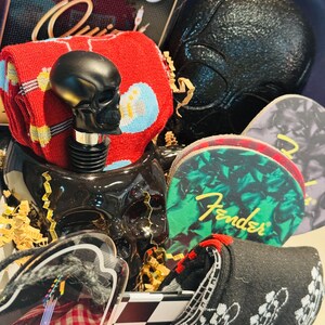 May include: A gift basket with a black skull bottle stopper, guitar picks, and socks. The socks are red with blue and yellow musical notes, and black with a guitar design. The guitar picks have the Fender logo.