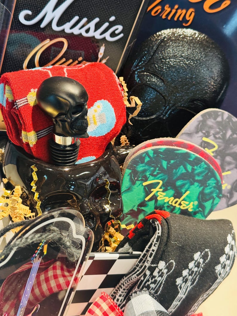 May include: A gift basket with a black skull bottle stopper, guitar picks, and socks. The socks are red with blue and yellow musical notes, and black with a guitar design. The guitar picks have the Fender logo.