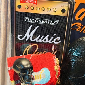 May include: A black skull bottle stopper with a red sock featuring a blue guitar design. The background includes a decorative box with the text "THE GREATEST Music" and a vintage amplifier design.